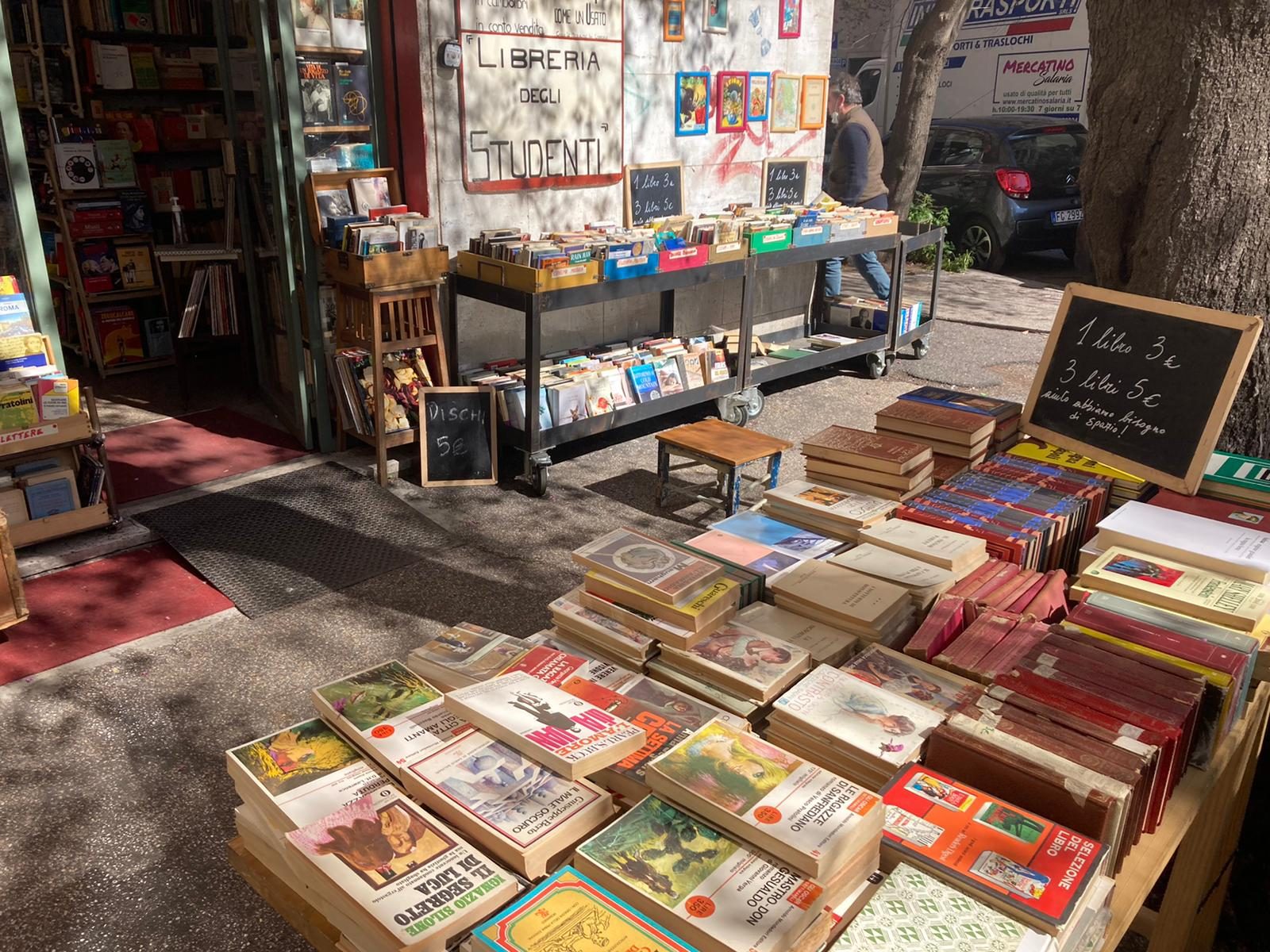Il marciapiede davanti alla libreria: carrelli e bancali di libri usati, un tavolo di legno e le lavagne con i prezzi scritti a gesso.